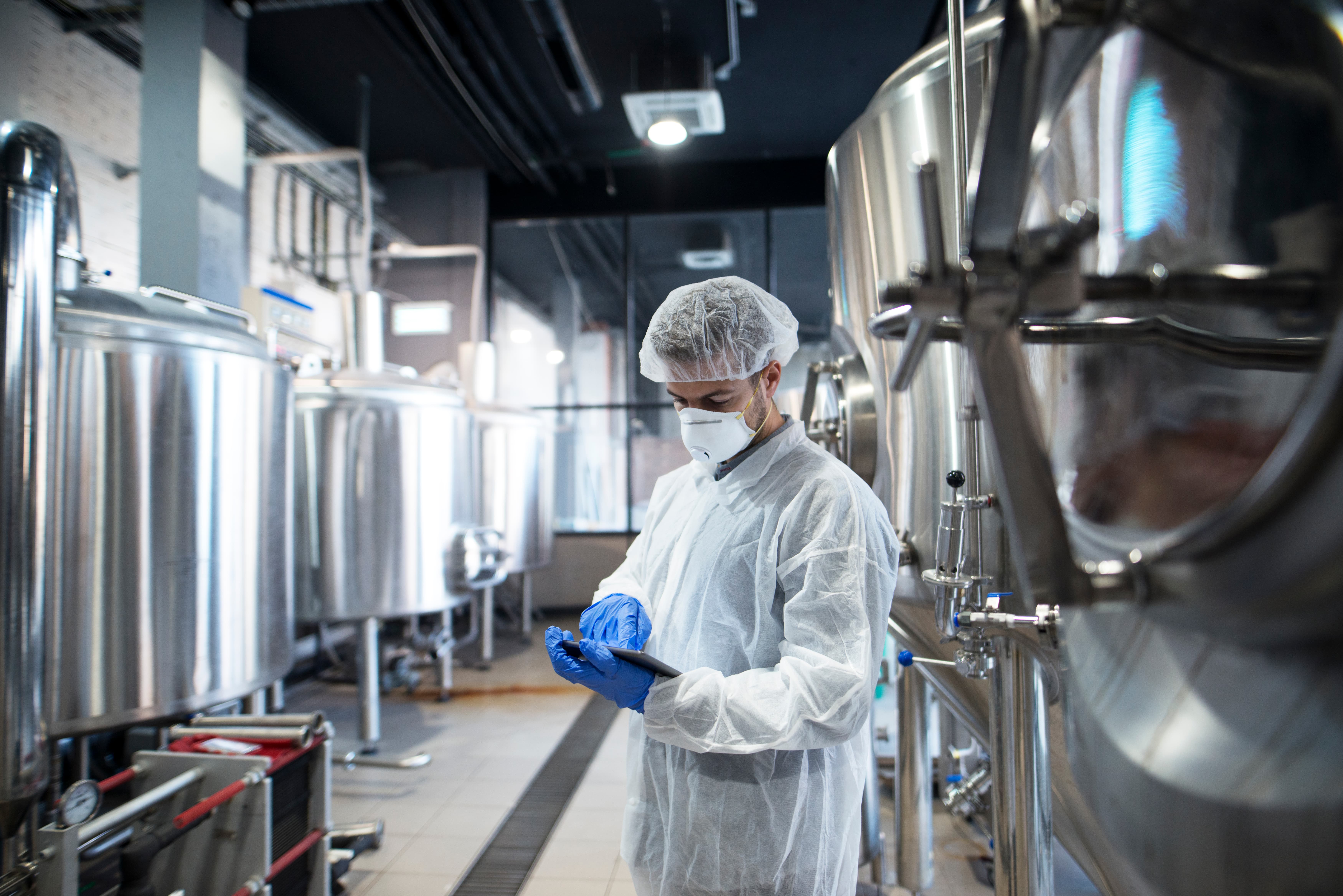 Food technologist in hygiene gear checking production data beside stainless steel process tanks